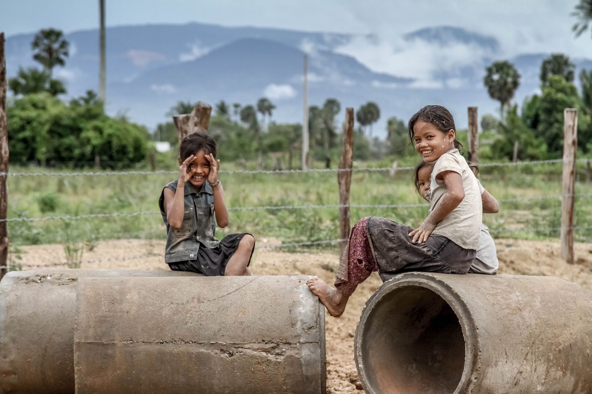 Two children on a concrete pipe at the edge of a rural road, hills behind.