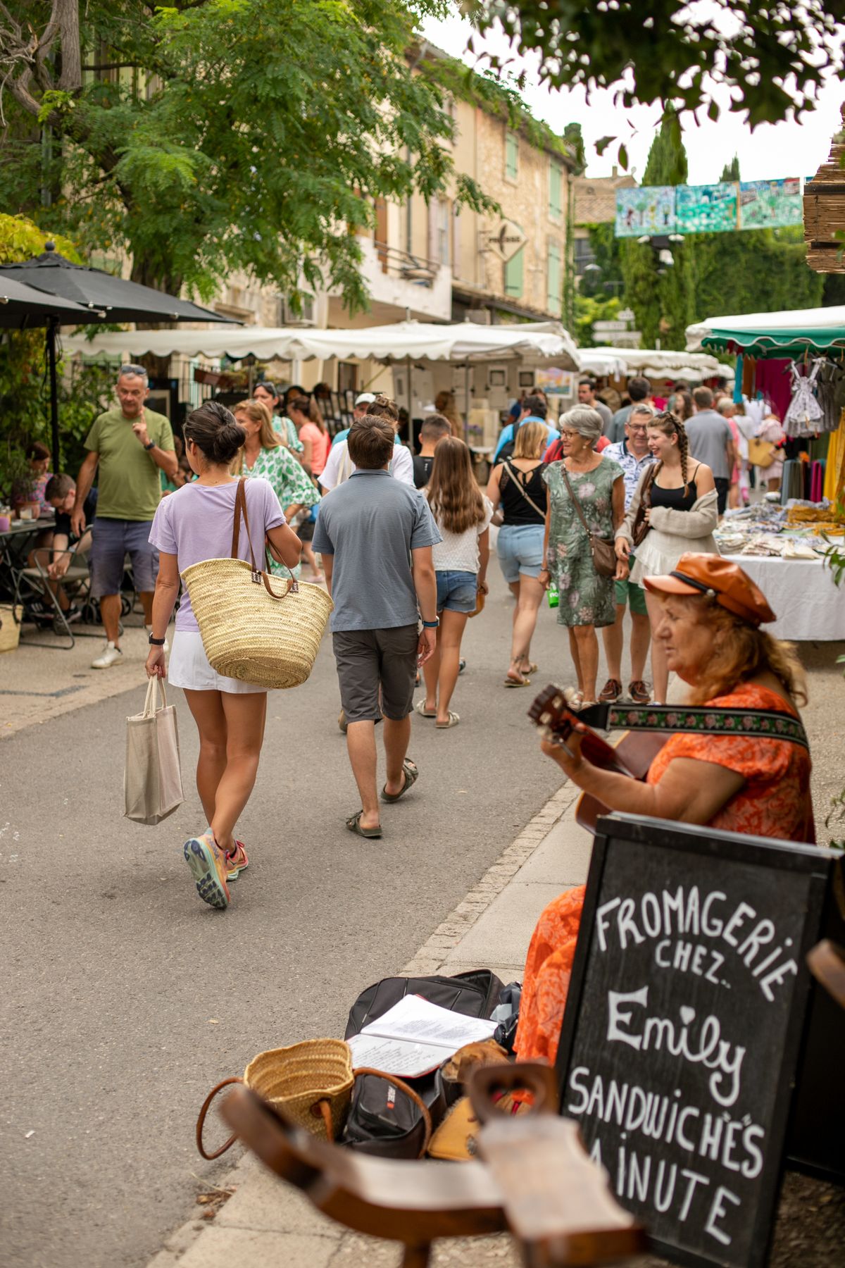 A Provencal market street under white awnings and plane trees, with shoppers carrying woven baskets