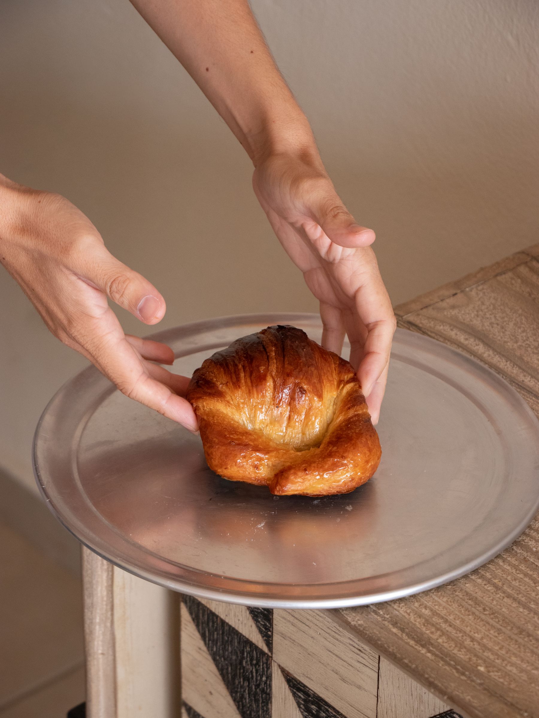 A warm golden medialuna pastry on a silver tray held in two hands